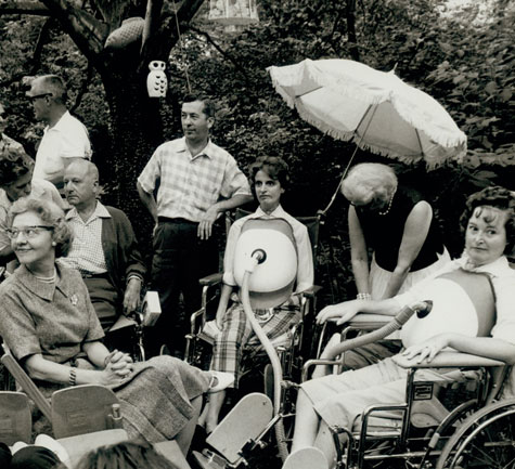 A gathering of 7 people in the backyard of Gini Laurie, several of whom are wearing turtle shells, mobile respiratory devices that attach to the upper abdomen to aid in breathing.